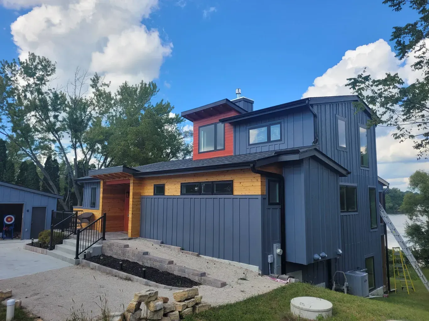 A modern two-story home with blue vertical siding, natural wood accents, a metal roof, and large windows near a lake.