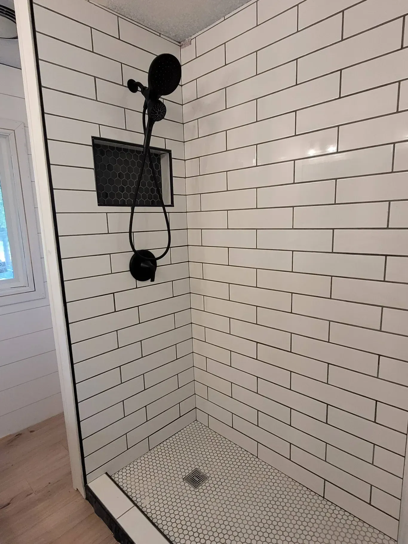 A walk-in shower with white subway tile walls, a black shower fixture, a recessed niche, and a small patterned tile floor.
