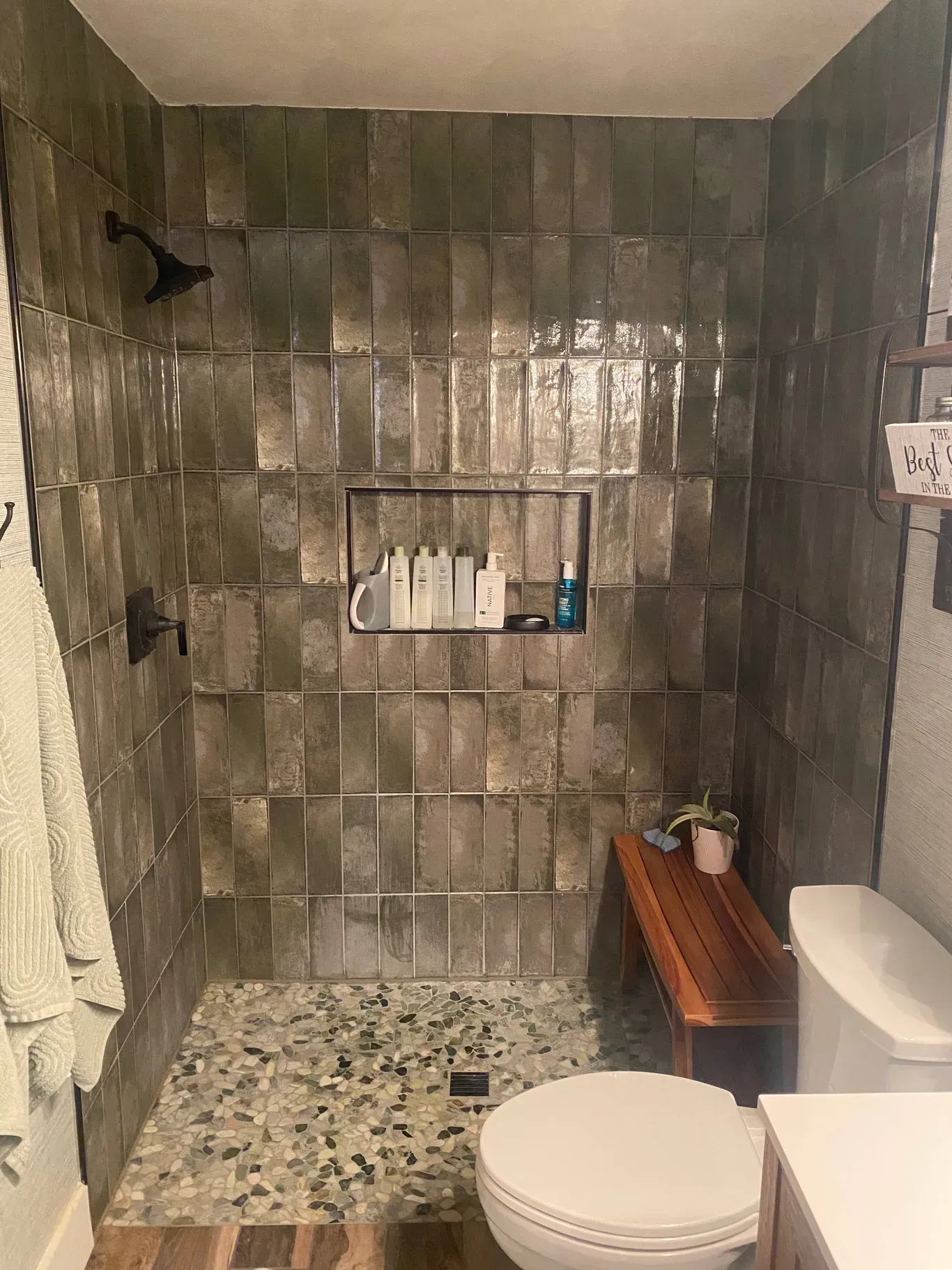 A walk-in shower with vertical, dark green tiles, a pebble floor, a built-in shelf, and a wood bench next to a toilet.