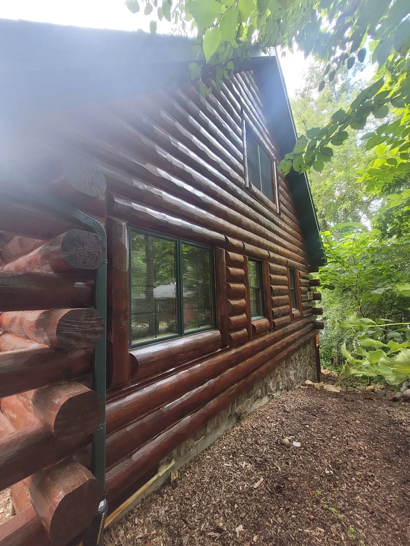 A brown log cabin exterior with windows and a stone foundation, viewed from the side amidst trees on a sunny day.