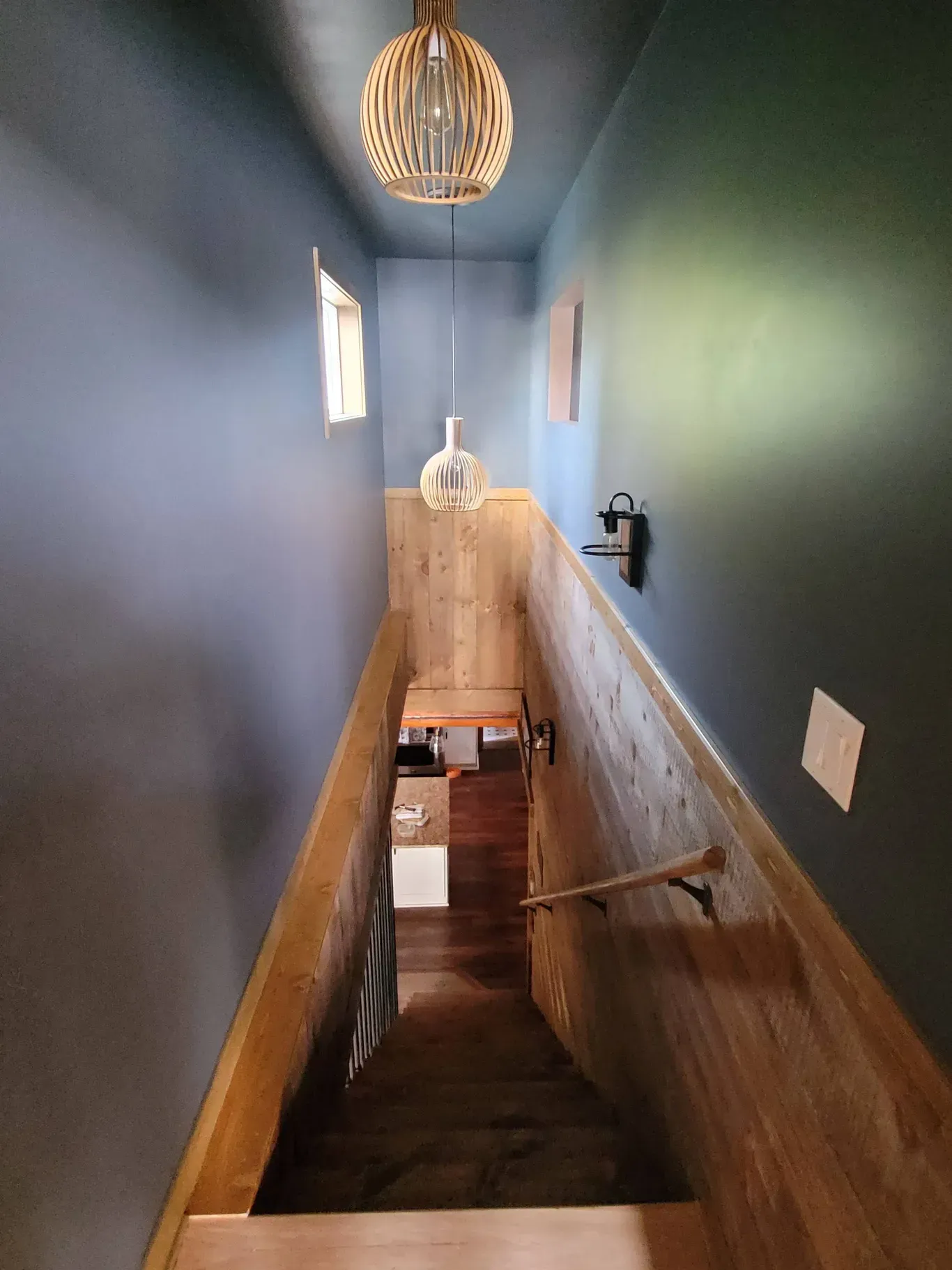 A high-angle view looking down a narrow staircase with dark blue walls, light wood paneling, and two hanging pendant lights.