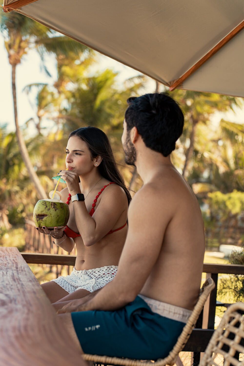 Um homem e uma mulher estão sentados sob um guarda-sol na praia bebendo água de coco.
