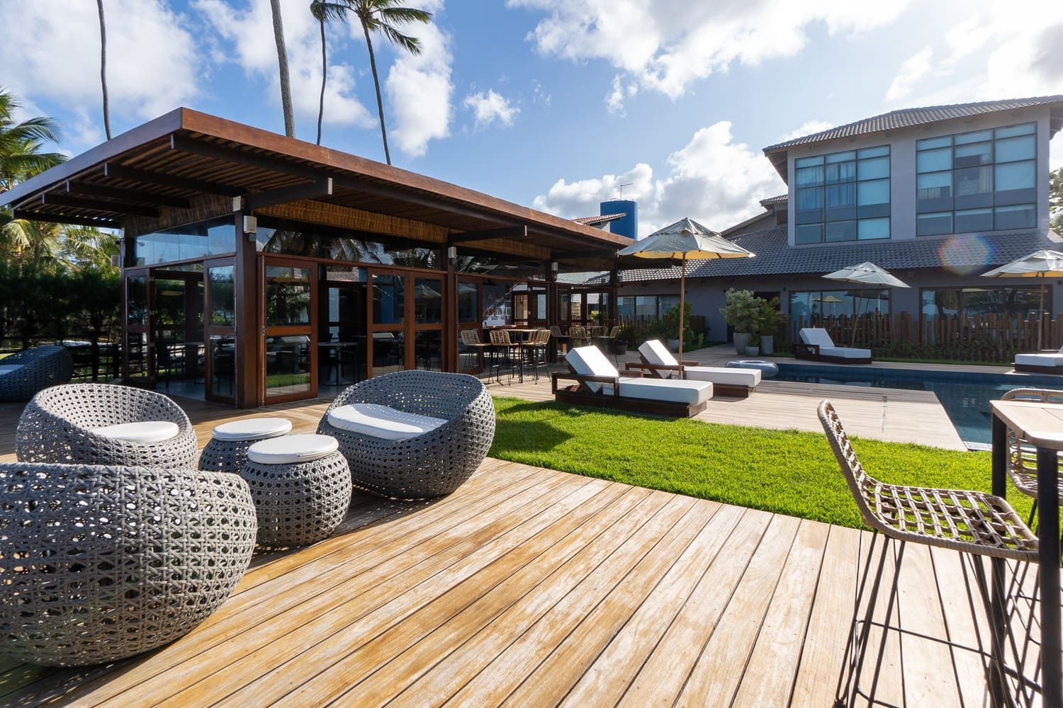 Um deck de madeira com móveis de vime e uma piscina ao fundo.