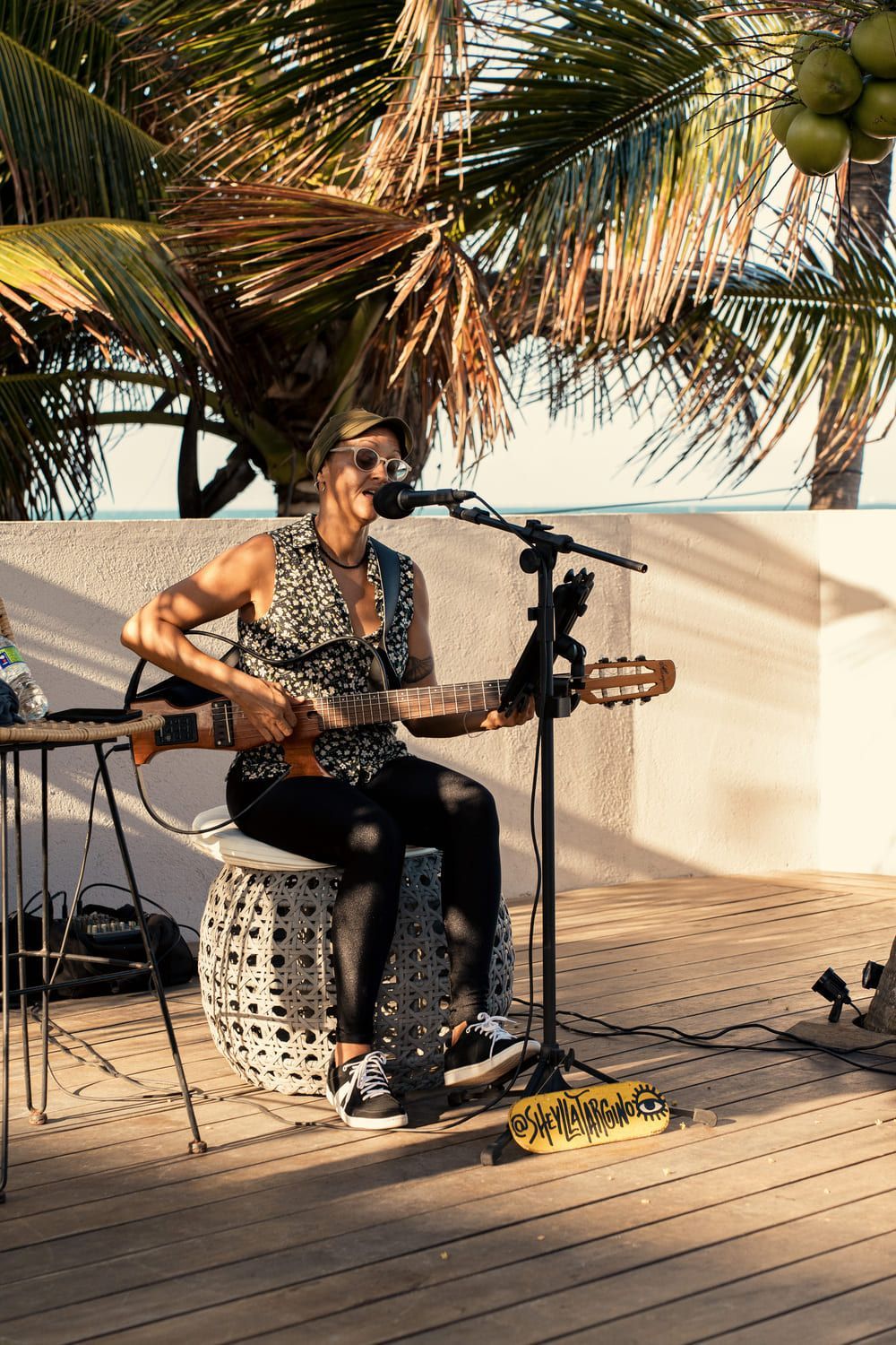 Uma mulher está sentada em um banquinho tocando violão e cantando em um microfone.