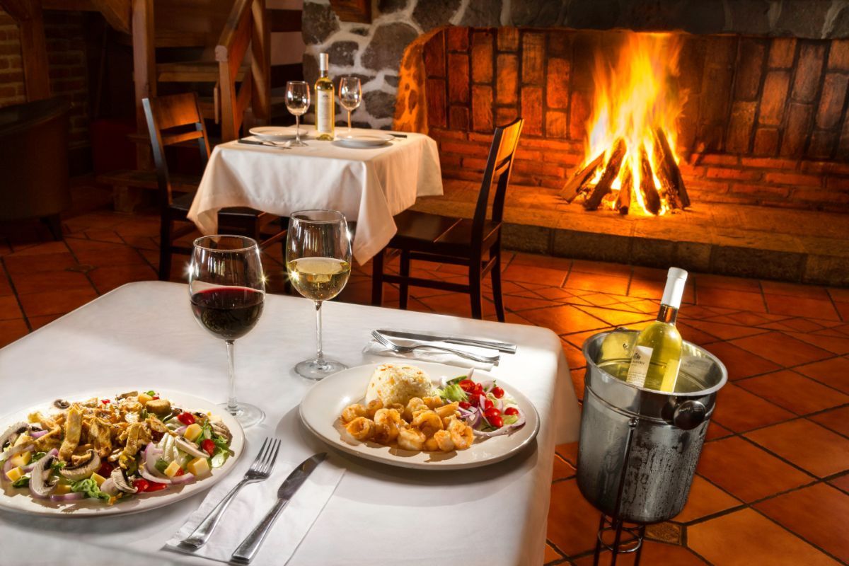 Una mesa con platos de comida y copas de vino frente a una chimenea.