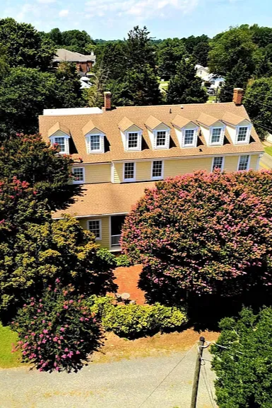 Yellow multi-story building with dormer windows, surrounded by green trees and flowering pink bushes.