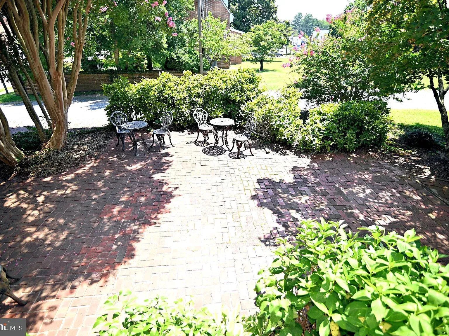 Brick patio with wrought iron table and chairs, surrounded by greenery and trees.