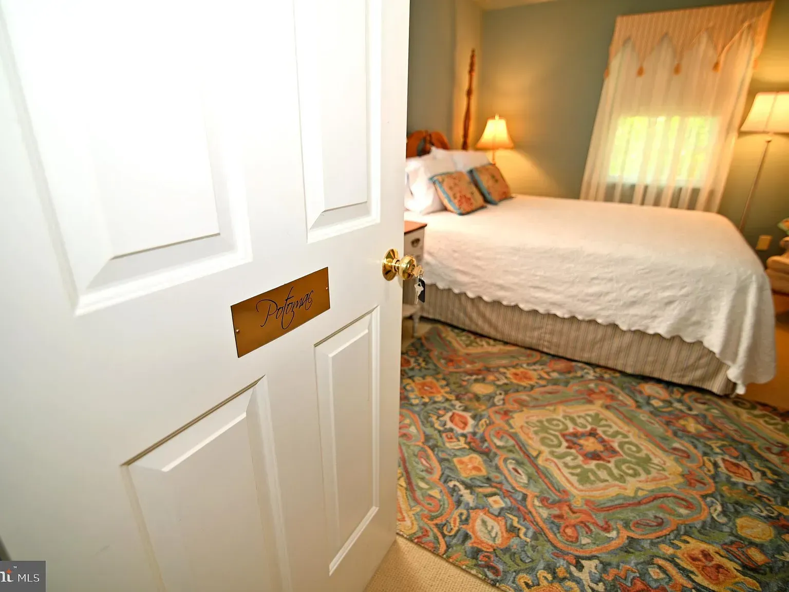Bedroom interior, open white door, bed with white bedding, patterned rug, lamp.