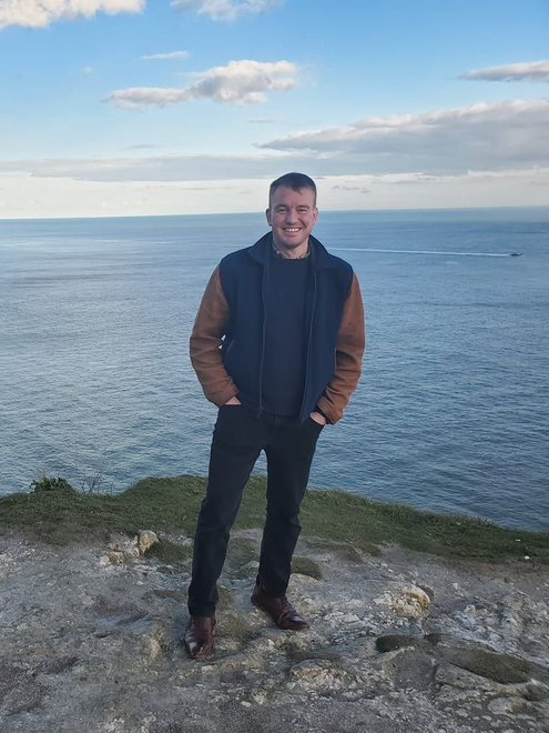 Man standing on cliff overlooking ocean, wearing jacket and dark pants. Cloudy sky.