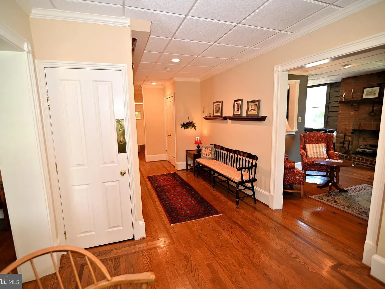 Hallway with wooden floor, white door, bench, and view into a living room with fireplace.