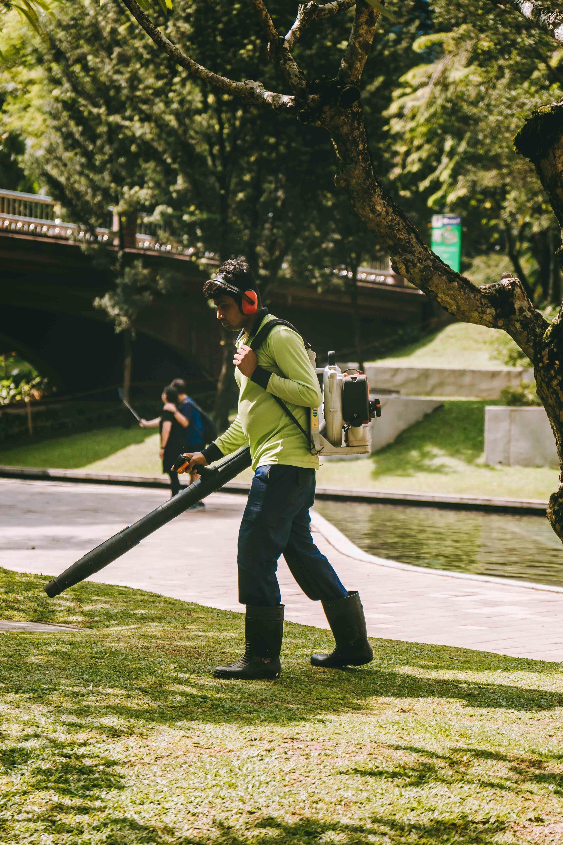 A man wearing safety equipment uses a leaf blower on a grassy area next to a river.