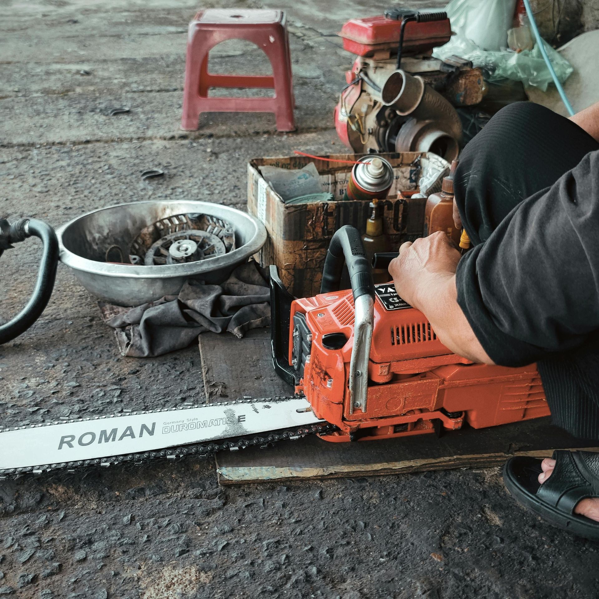 Person works on an orange chainsaw on a concrete surface. Tools, engine, and red stool in background.