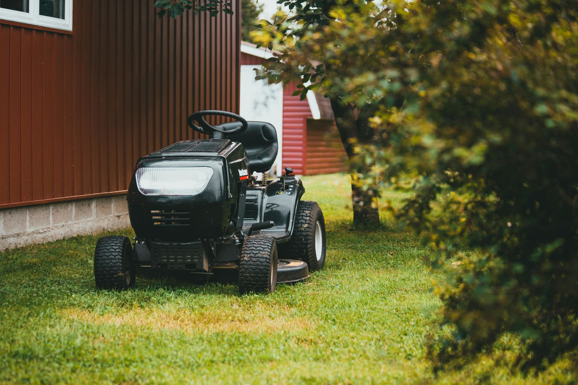 Black riding lawn mower on green grass, near a red building.