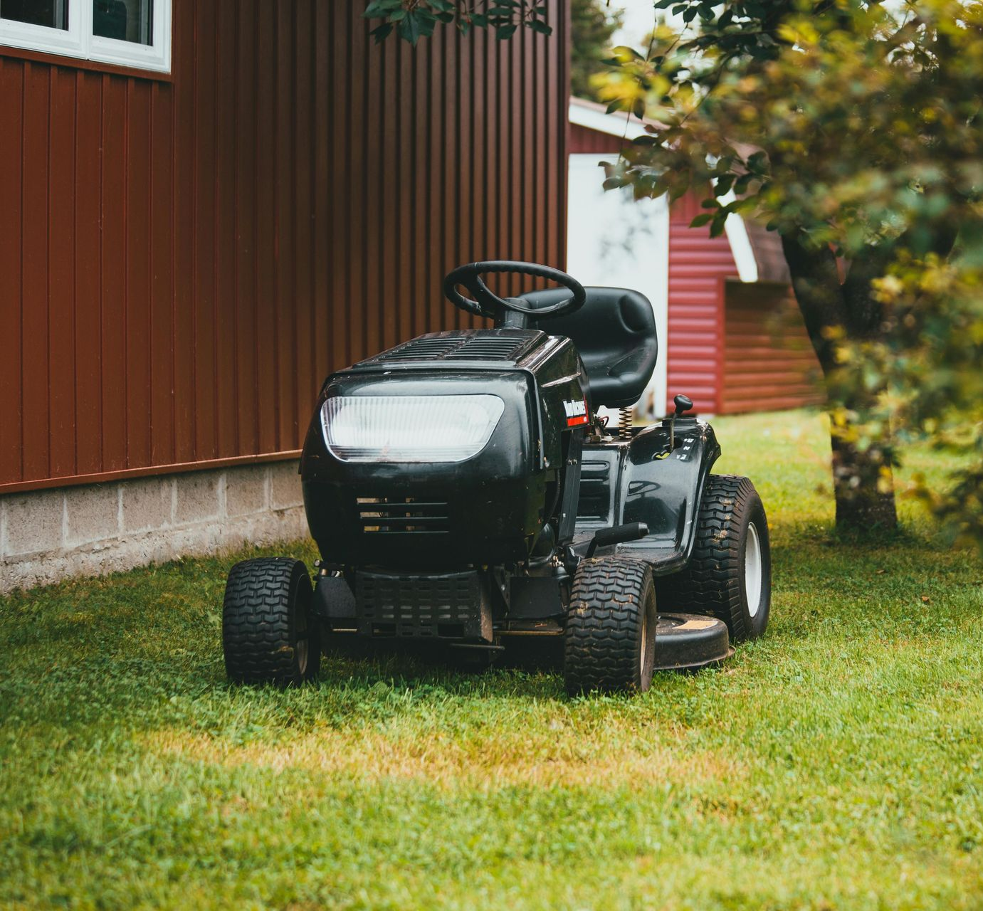 Black riding lawnmower parked on green grass beside a brown building with a white window.