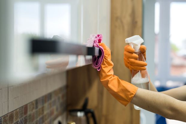 Person wearing orange gloves cleaning a kitchen cabinet with spray bottle and pink cloth.