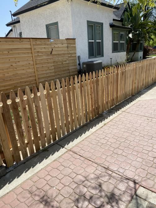 A wooden picket fence surrounds a white house on a brick sidewalk.