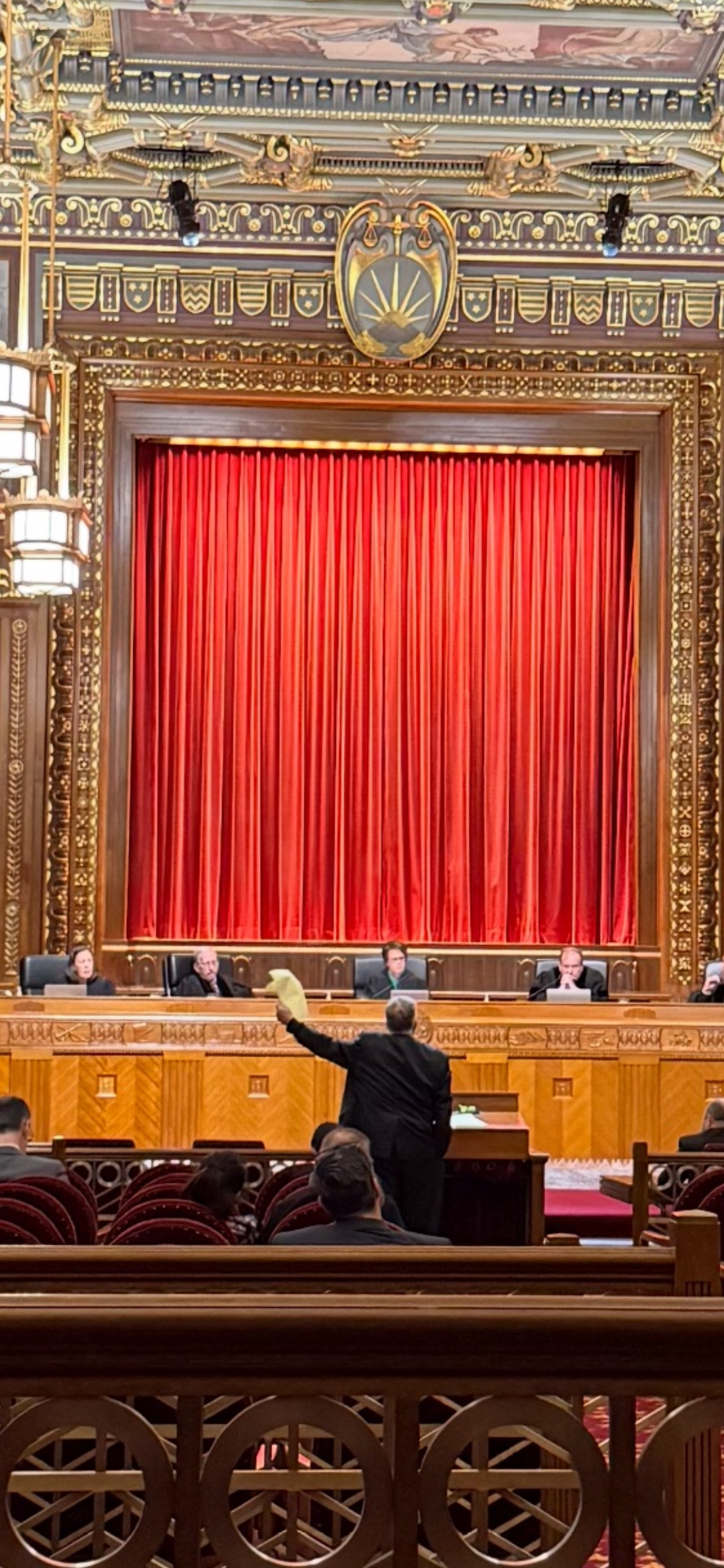 Inside a courtroom, a man gestures toward a red curtain behind a bench. 