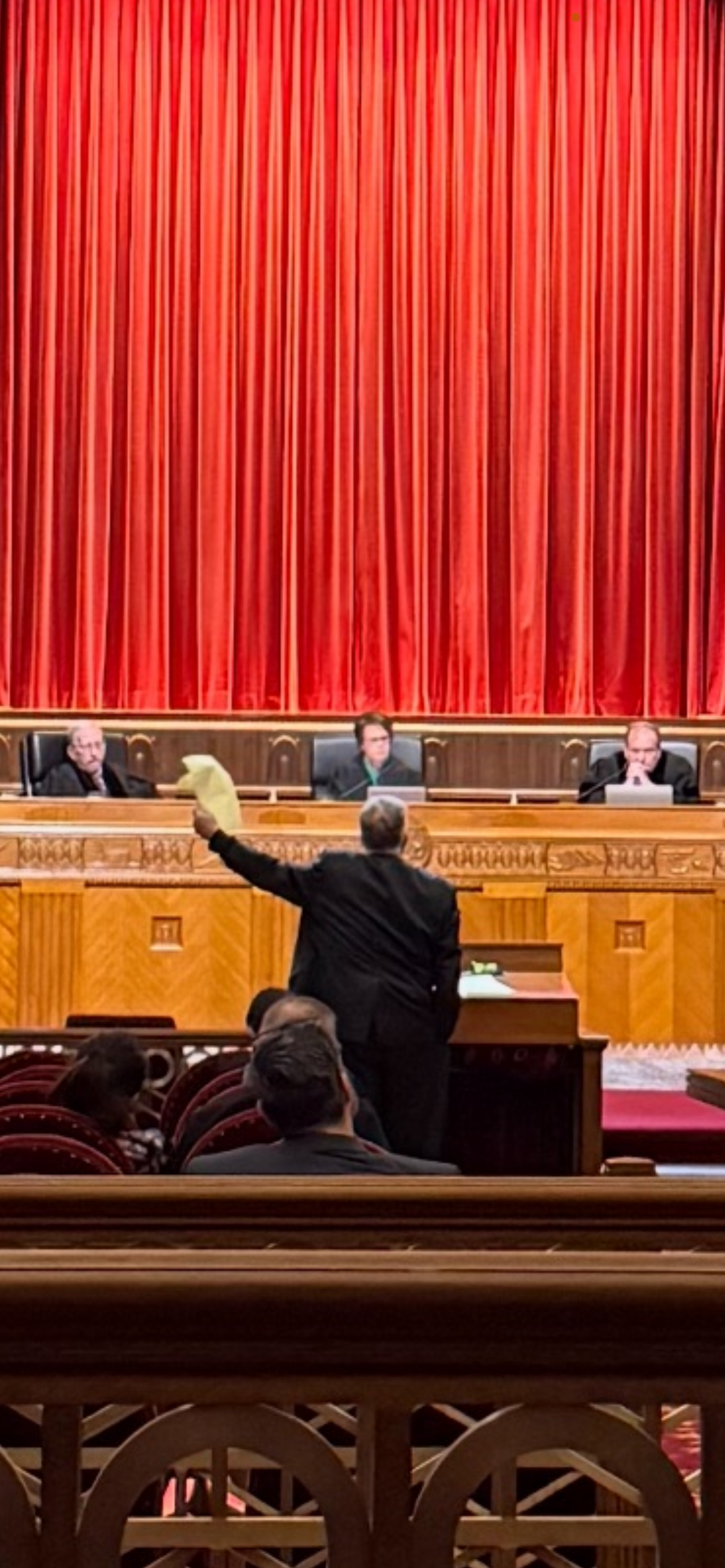 A person in a courtroom raising a paper. Judges sit in the background under a red curtain.