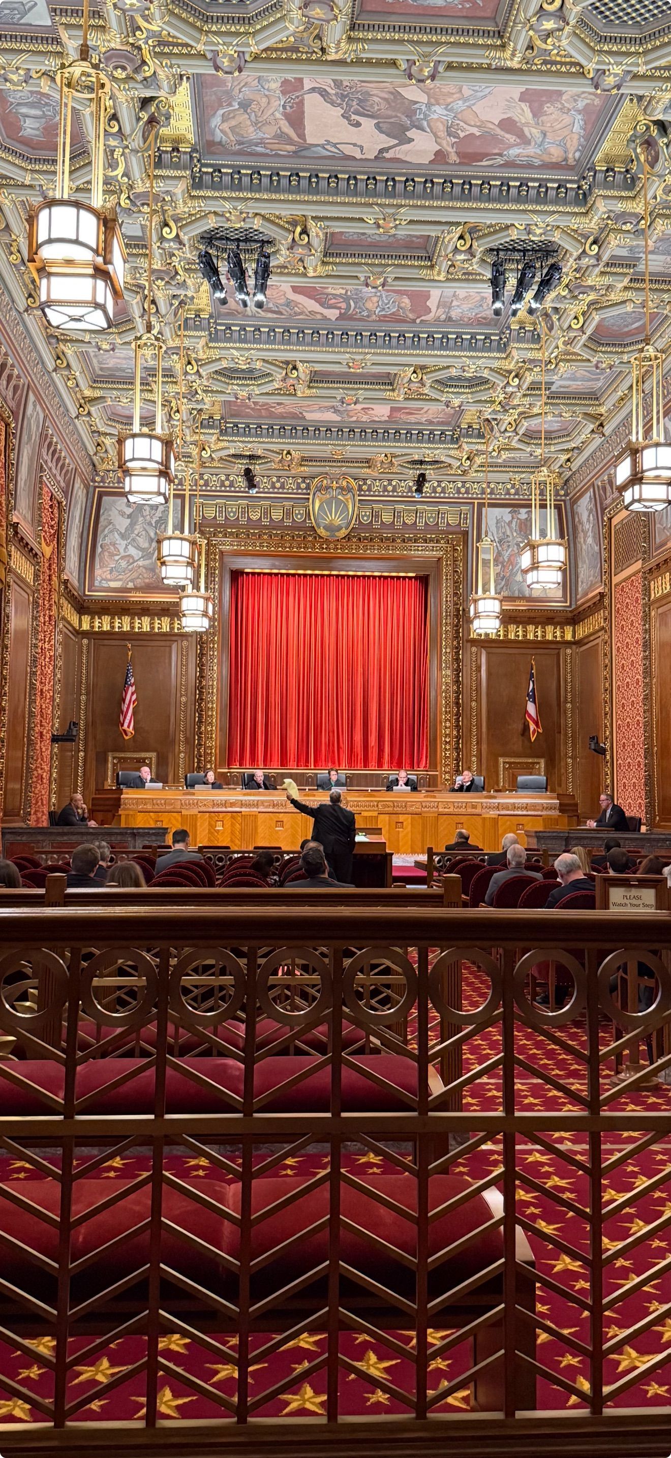 Ornate interior with a person addressing a crowd in front of a red curtain.