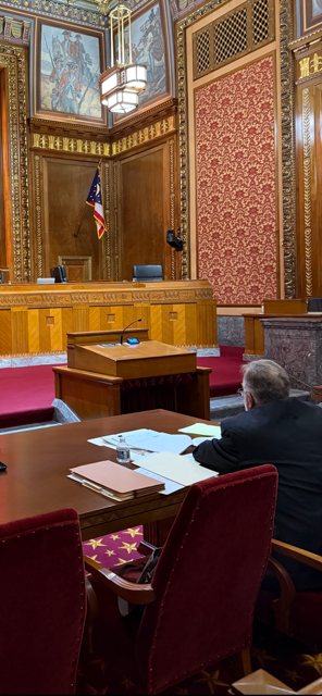 Interior of a courtroom with a person at a table, papers in front of them, facing a judge's bench.