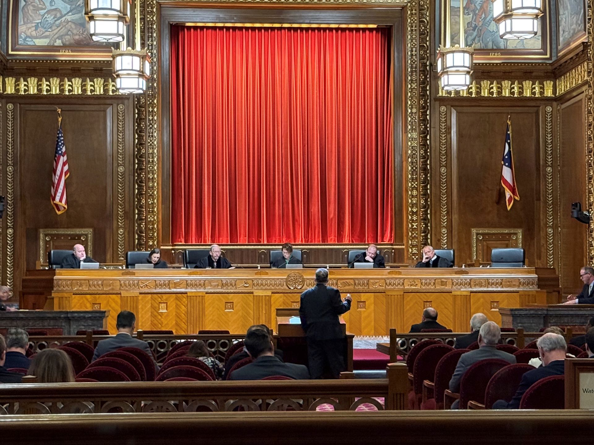 Courtroom with judges seated on a raised bench, a speaker at a podium, and an audience.