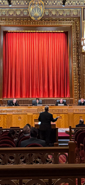 Ornate interior with a person addressing a crowd in front of a red curtain.
