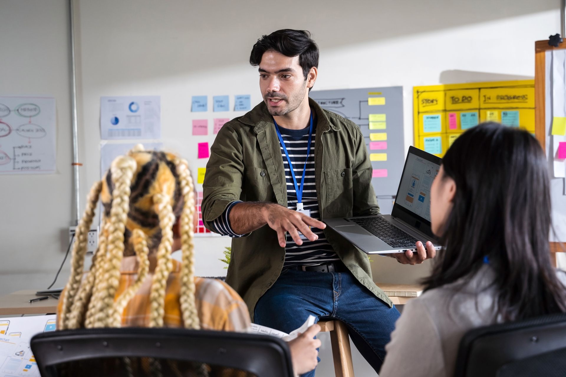A person in an olive jacket presents work on a laptop to two colleagues in an office with notes on the wall.