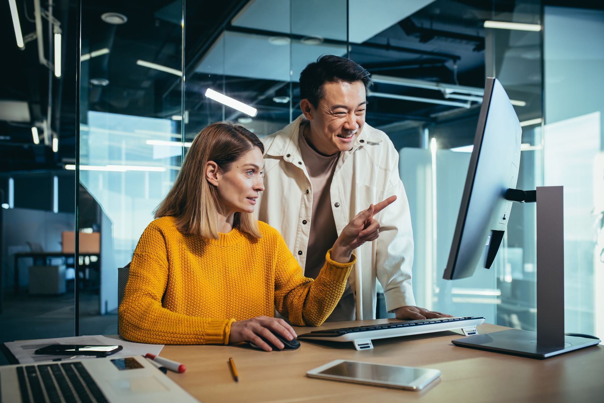 A woman in a yellow sweater and a man stand at a desk in a modern office, looking at a computer screen and collaborating.