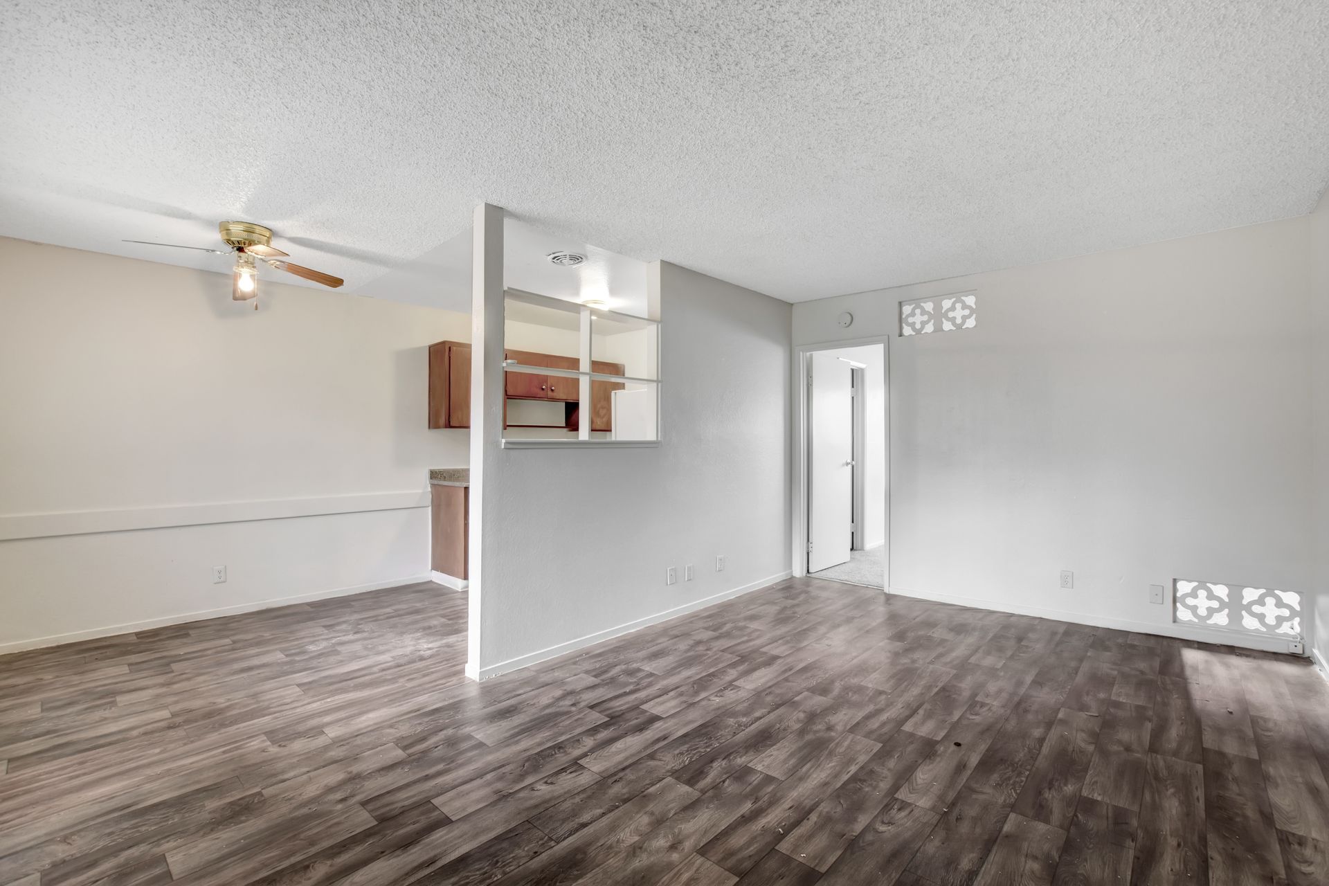 Empty gray living room with wood-look flooring, ceiling fan, and open pass-through to the kitchen.