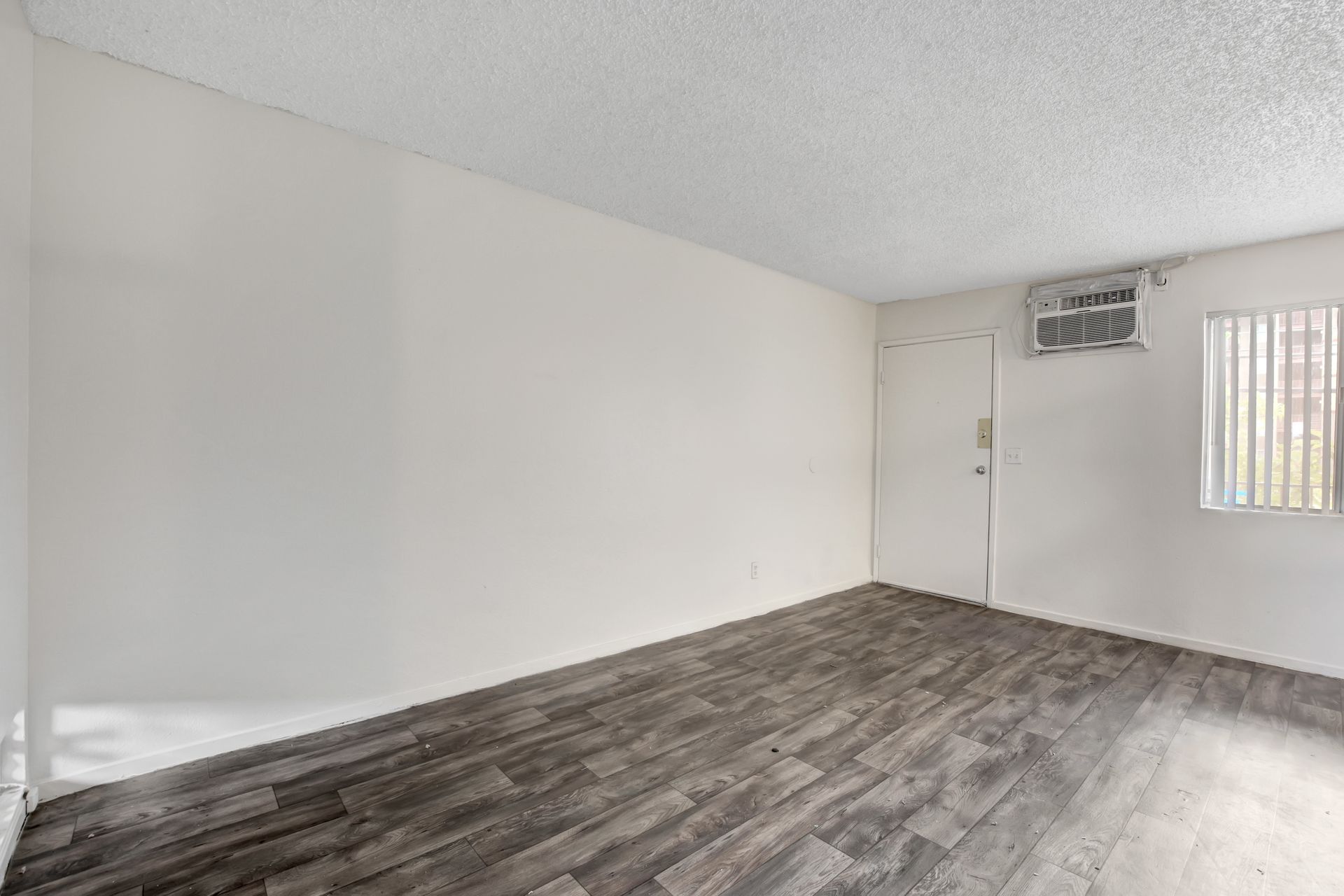 Empty room with white walls, gray wood flooring, small window, and wall-mounted air conditioner.