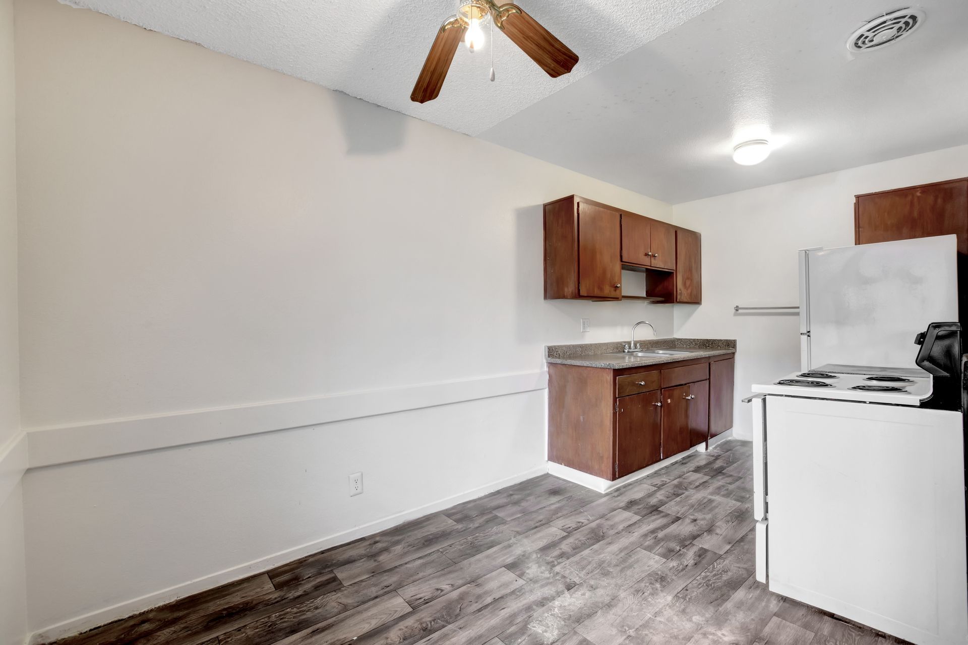 Small kitchen with white appliances, wood cabinets, gray flooring, and a ceiling fan in a bright room