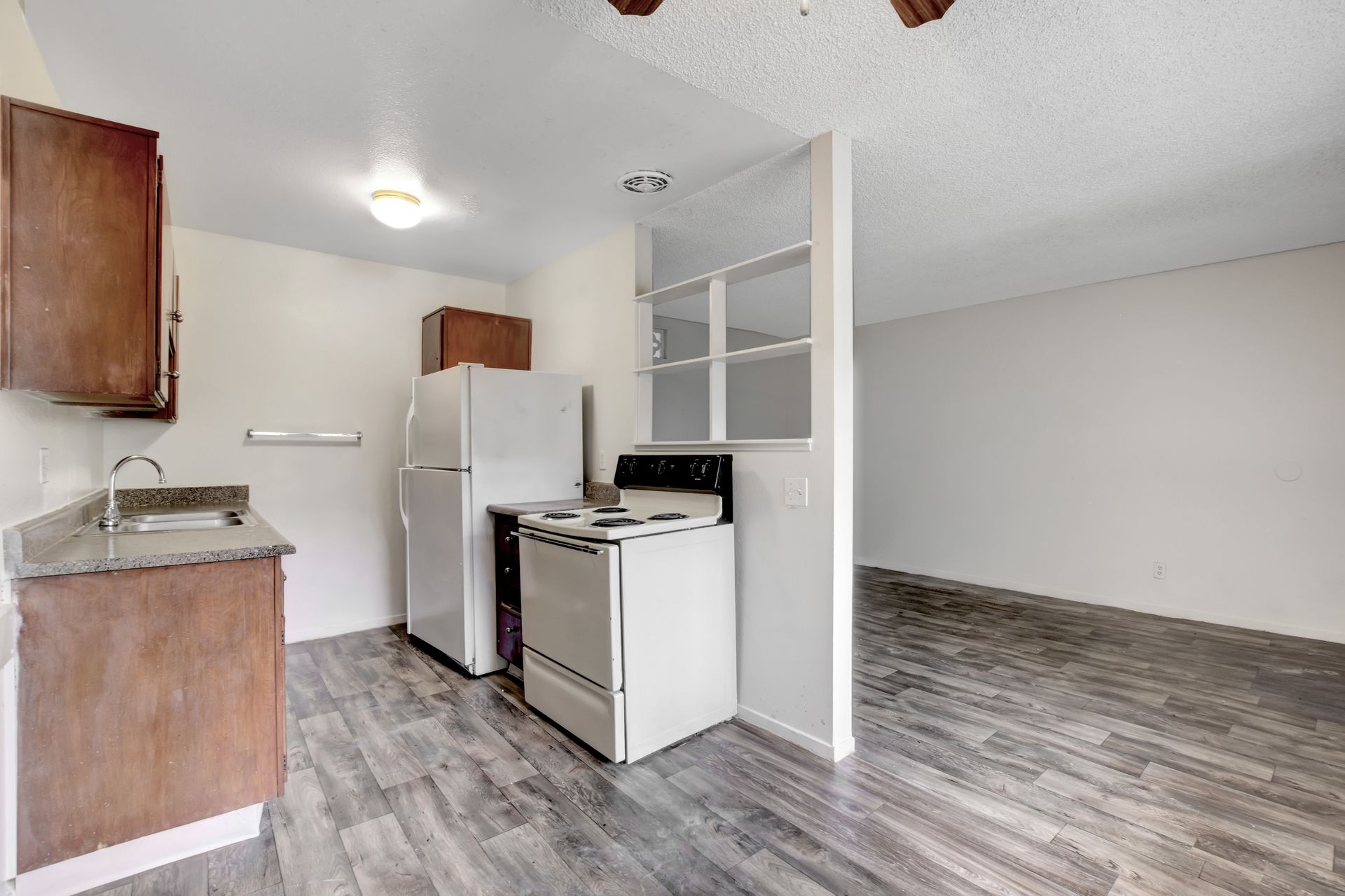Small kitchen with white appliances, wood cabinets, and open living area with gray flooring