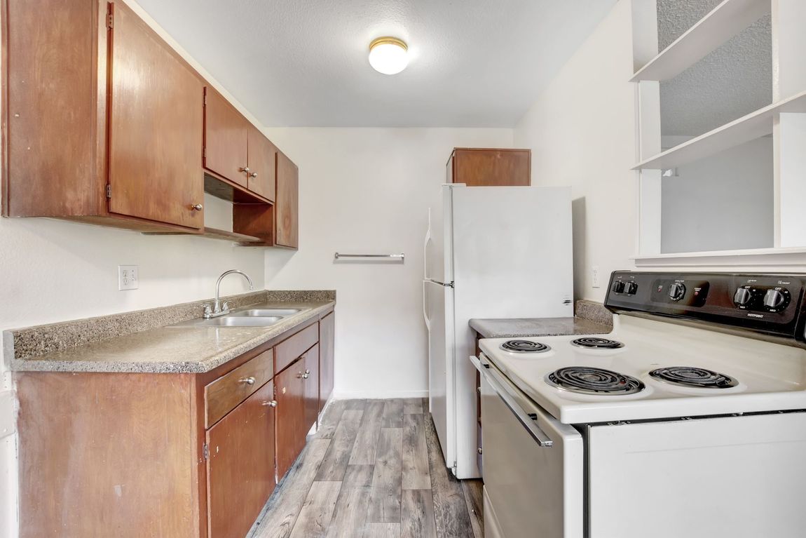 Narrow galley kitchen with wood cabinets, white appliances, and gray wood-look flooring.