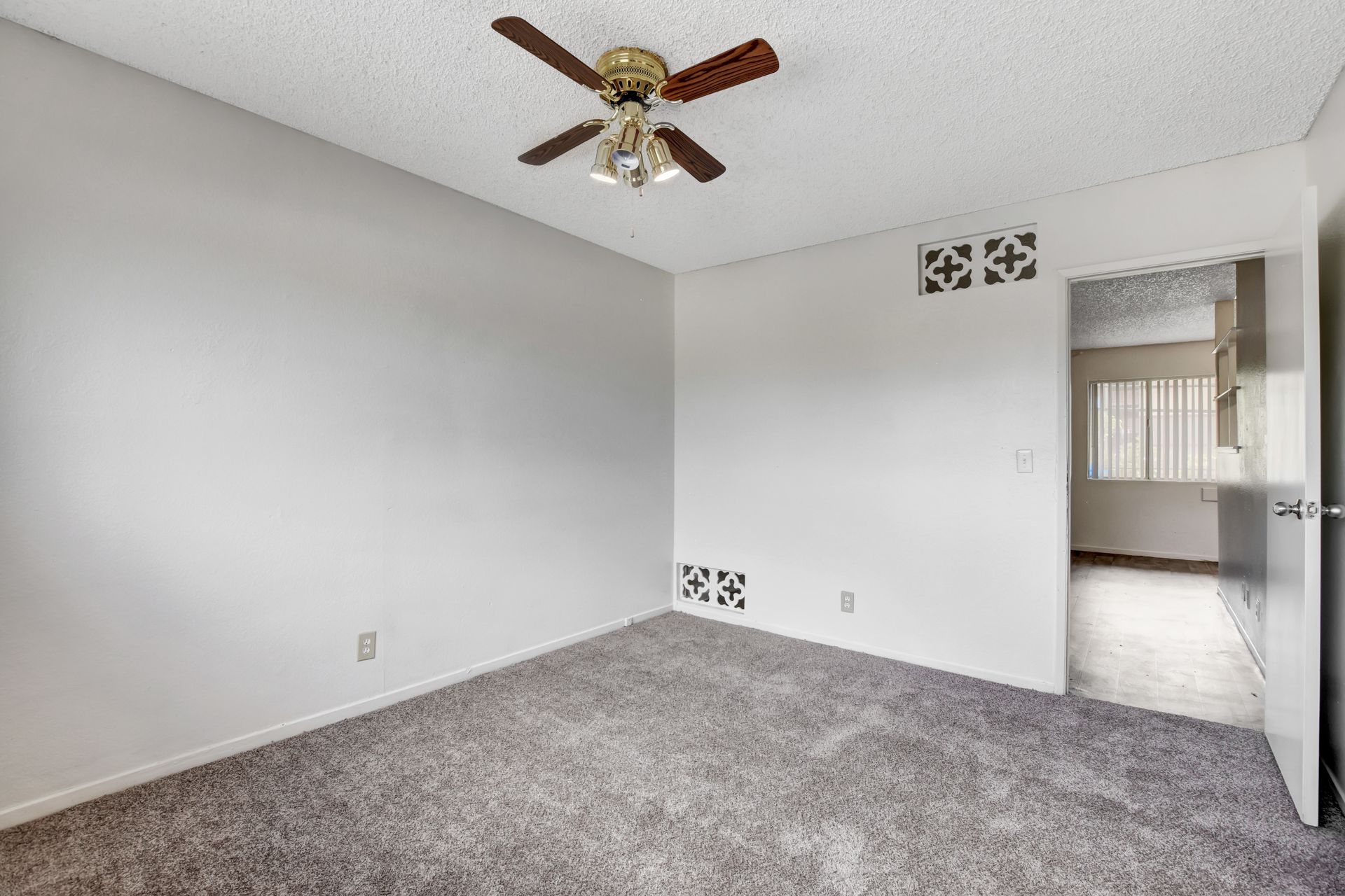 Empty carpeted bedroom with white walls, ceiling fan, and doorway to a bright adjoining room