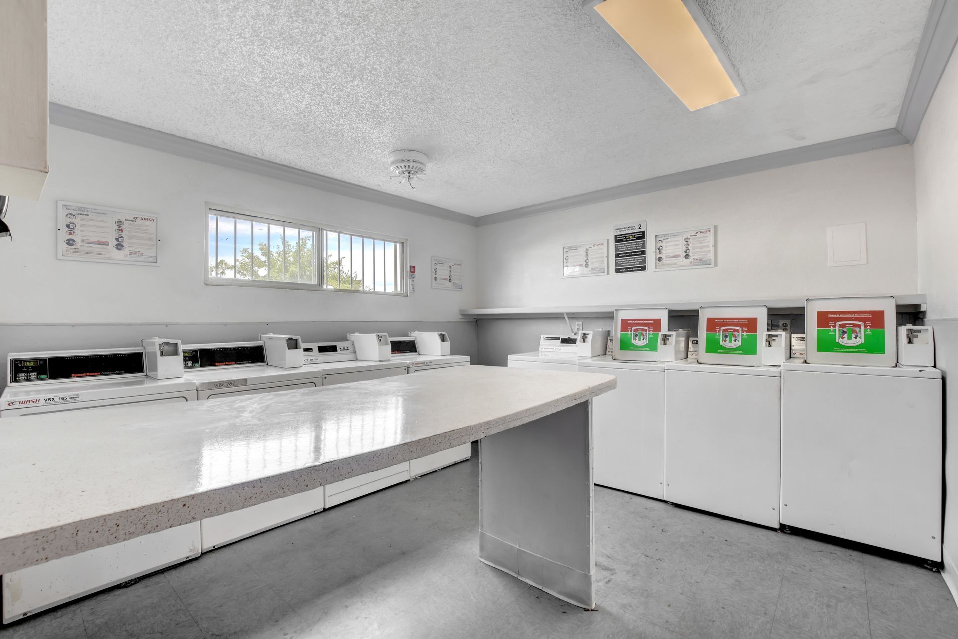 Bright laundry room with white folding tables, washers, dryers, and a windowed wall