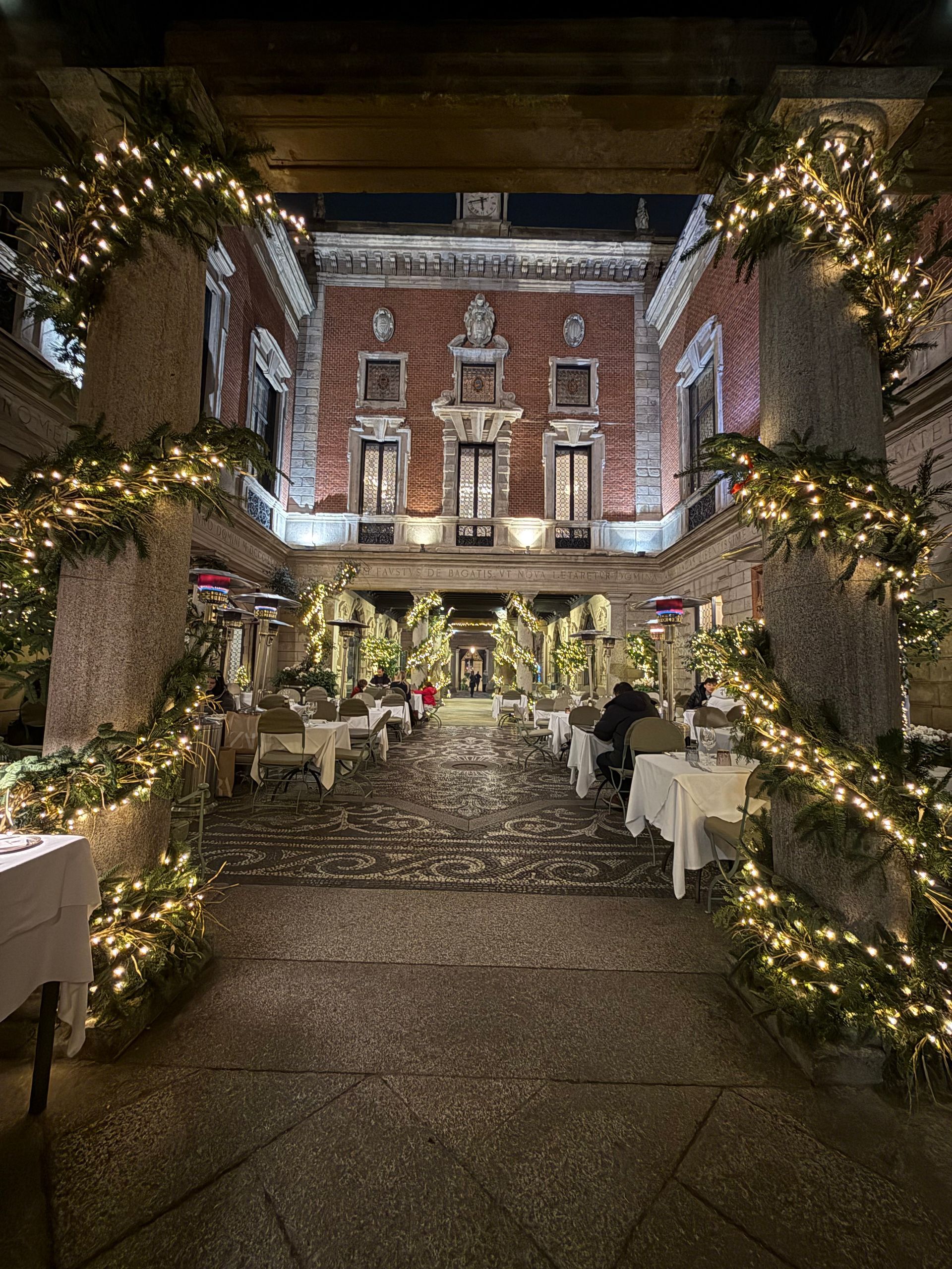 A large building is decorated with christmas lights and tables and chairs.