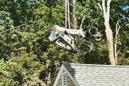 a bulldozer is hanging from a crane over a house .