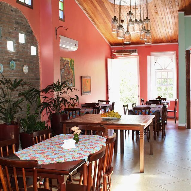 Interior de restaurante com mesas e cadeiras, paredes cor-de-rosa, plantas e luminárias pendentes.