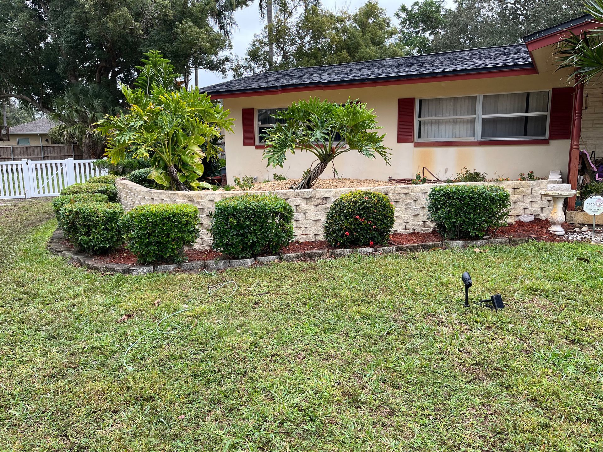 Bungalow with manicured lawn and hedge-lined garden bed. White fence on the left, trees in the background.