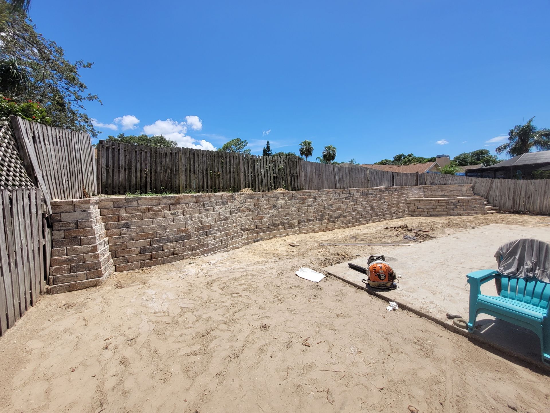 Backyard with a stone retaining wall, wooden fence, and a blue chair on a sunny day.
