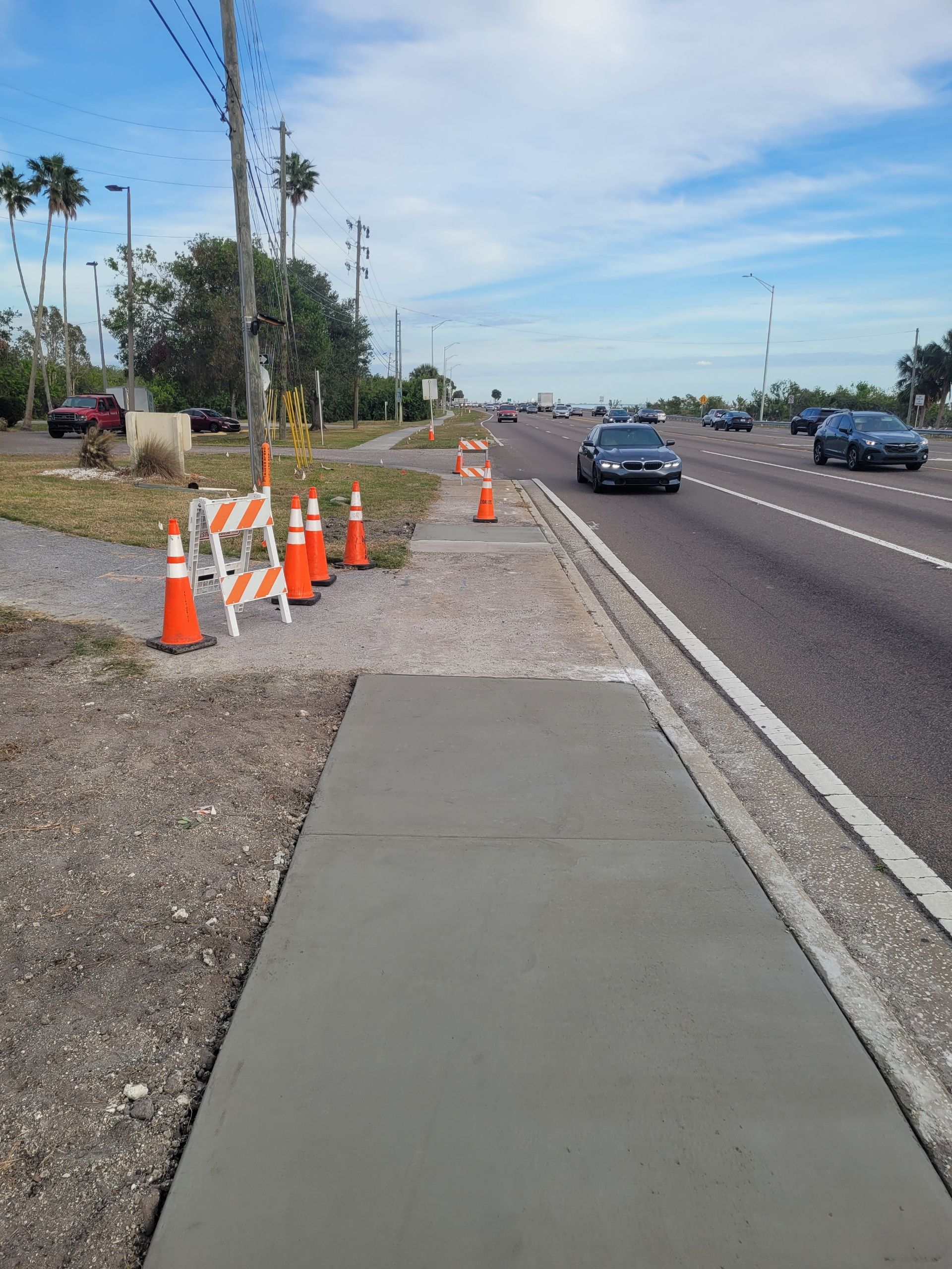 Newly poured concrete sidewalk next to a road, with orange cones and safety barricades.