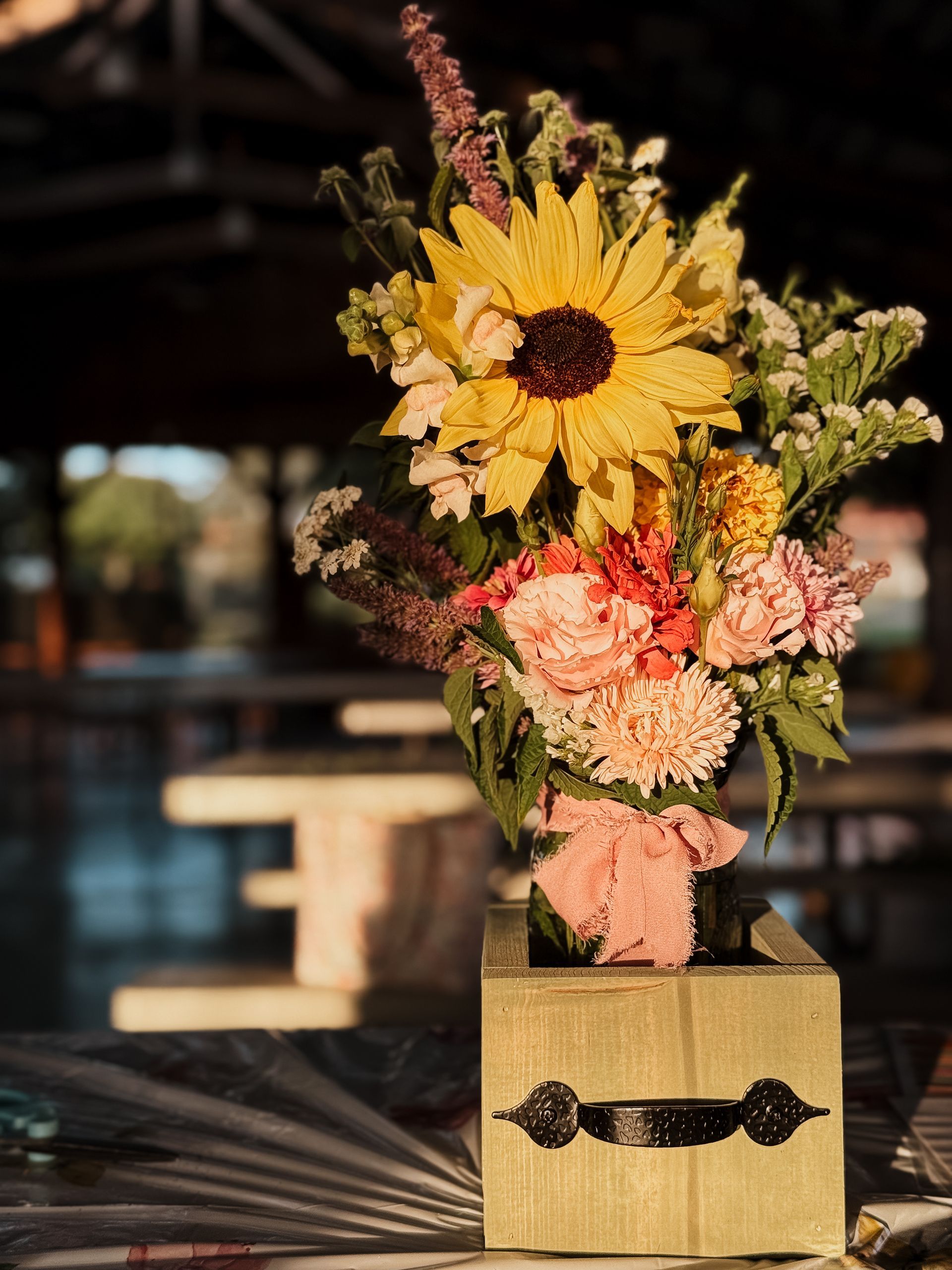 Sunflower and mixed flowers in a wooden box on a table. Blurred background, tables, and outdoor setting.