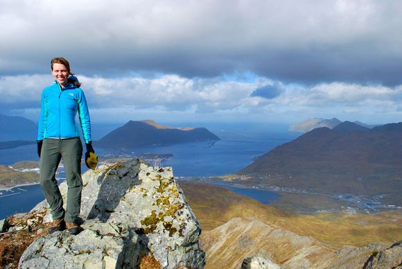 Person in a blue jacket stands on a rocky peak overlooking a lake and mountains under cloudy skies