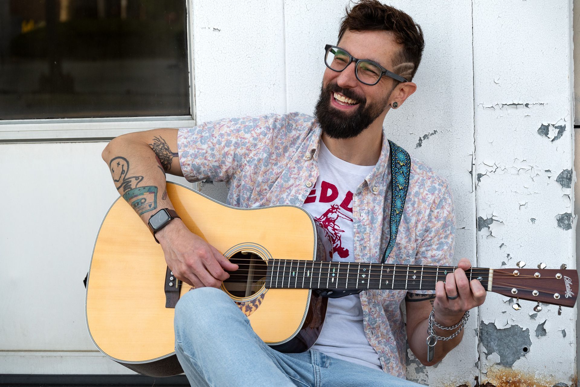 A smiling man with glasses and a beard plays an acoustic guitar, seated against a white wall. He wears a patterned shirt and jeans.