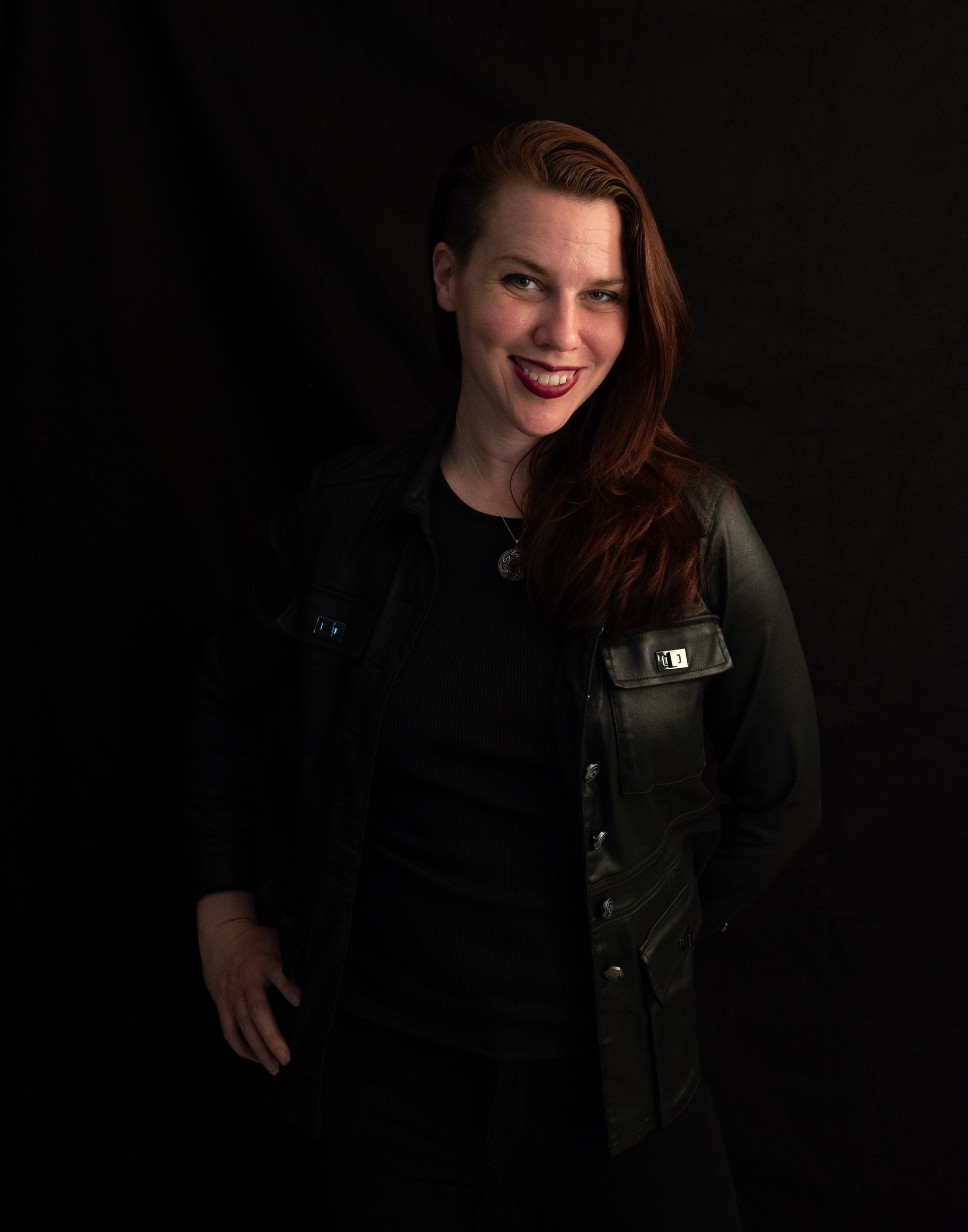 Woman with red hair and a leather jacket smiles, standing in front of a black background.