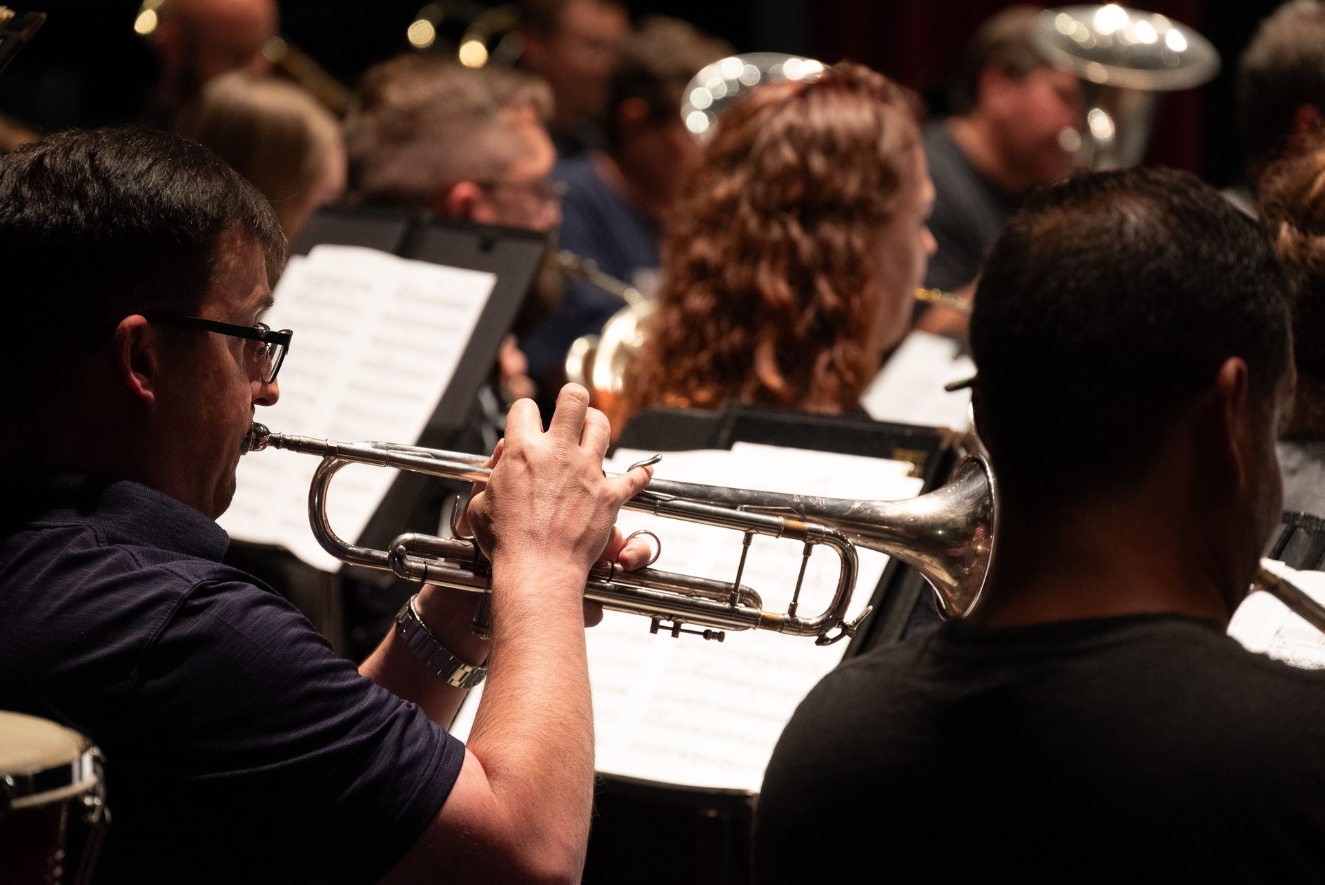 A musician in an orchestra playing a silver trumpet. He wears glasses and a dark blue shirt. Other musicians are visible in the background.