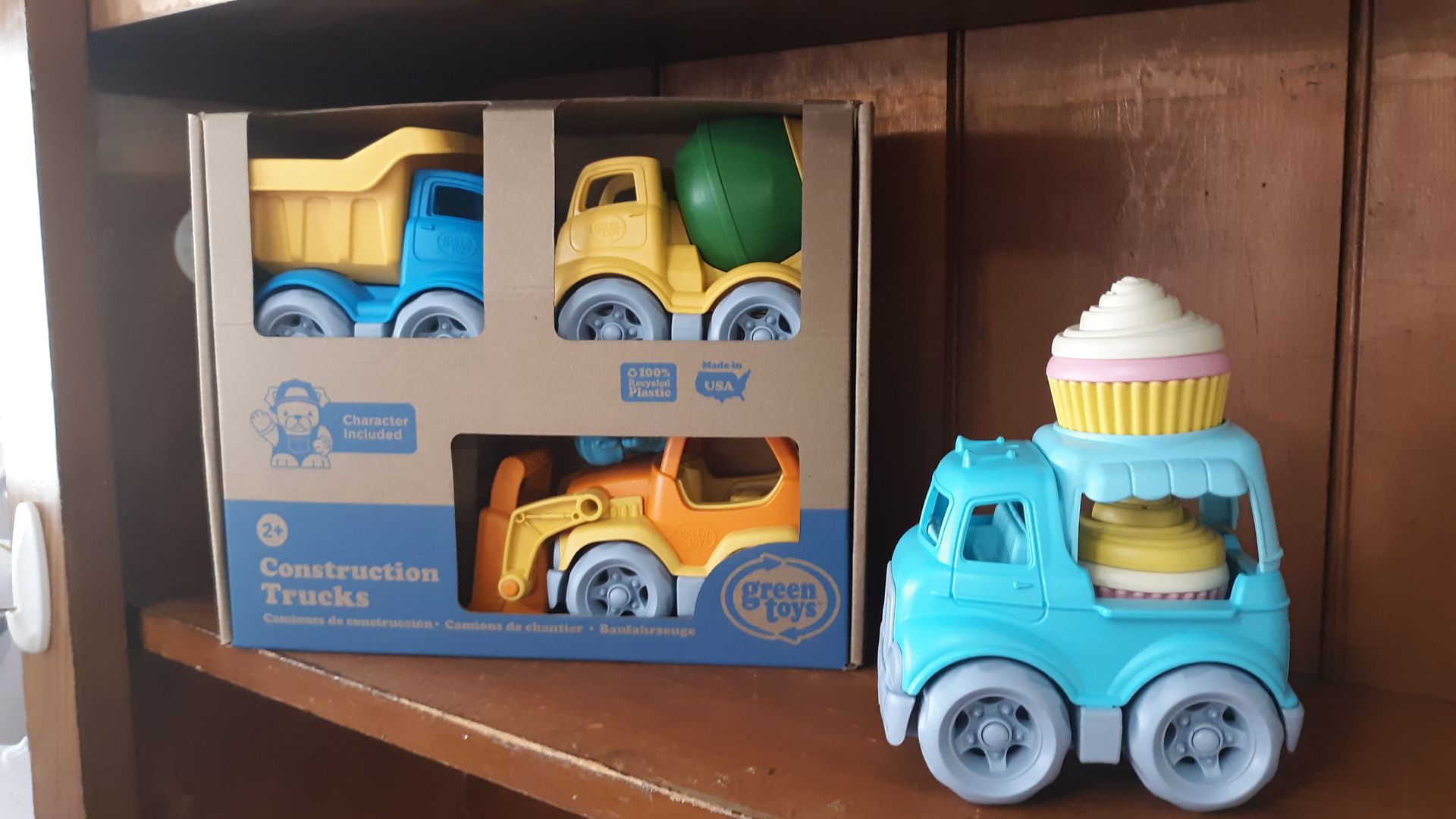 A box of construction trucks and an ice cream truck are sitting on a wooden shelf.