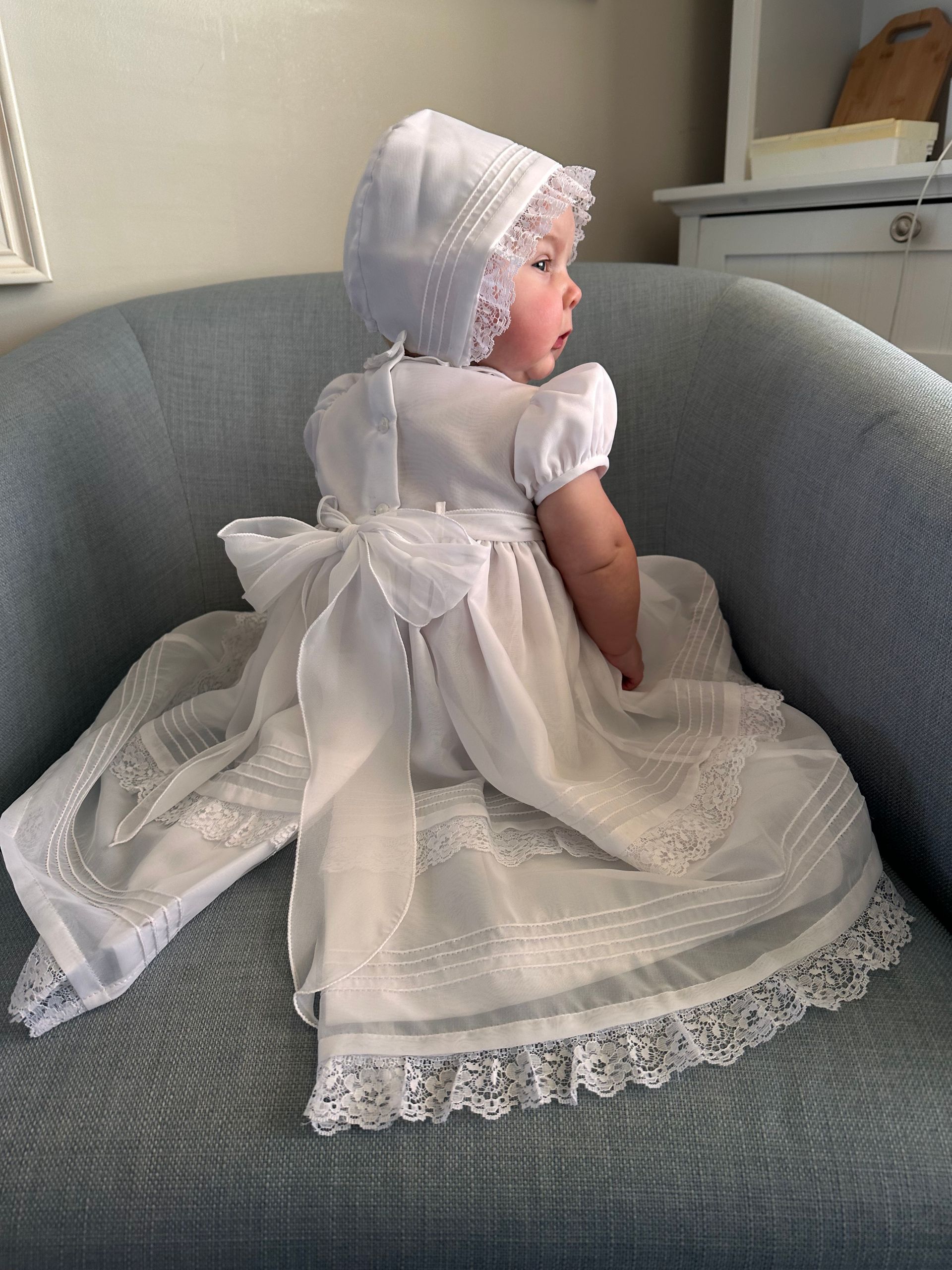 A baby in a white dress and bonnet is sitting on a chair.