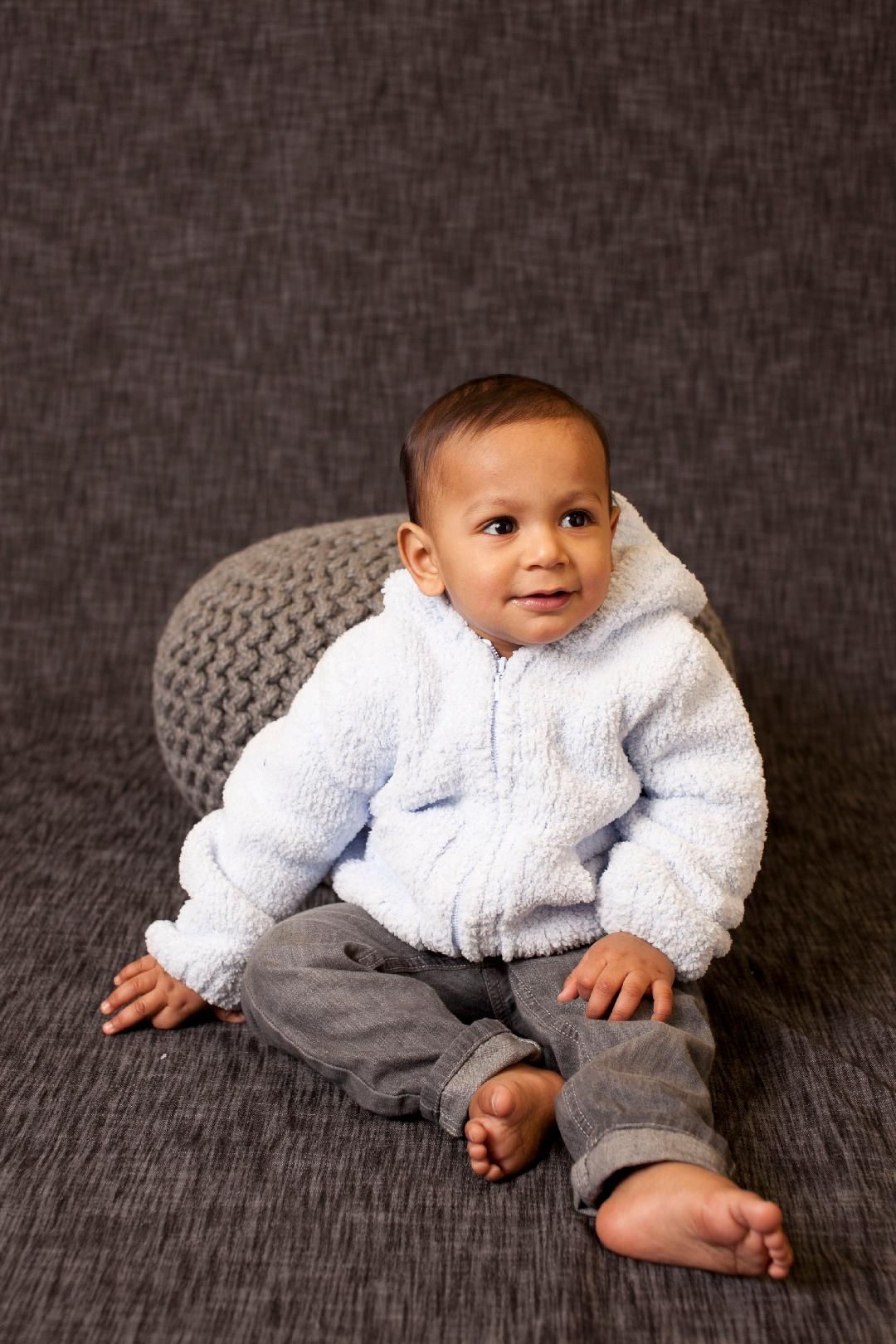 A baby is sitting on a couch wearing a white jacket and gray pants.