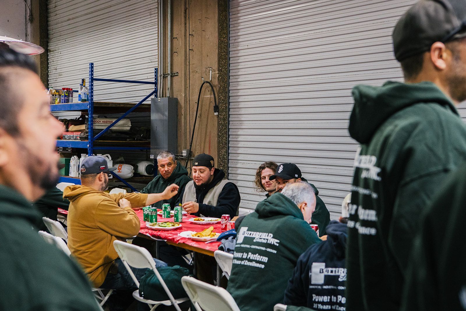 a group of men are sitting at a table in a garage eating food .
